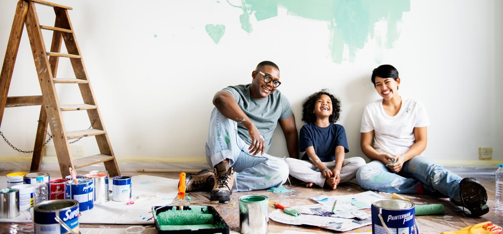 Young family sitting on the floor with a half-painted wall behind them. There are paint supplies on the floor.